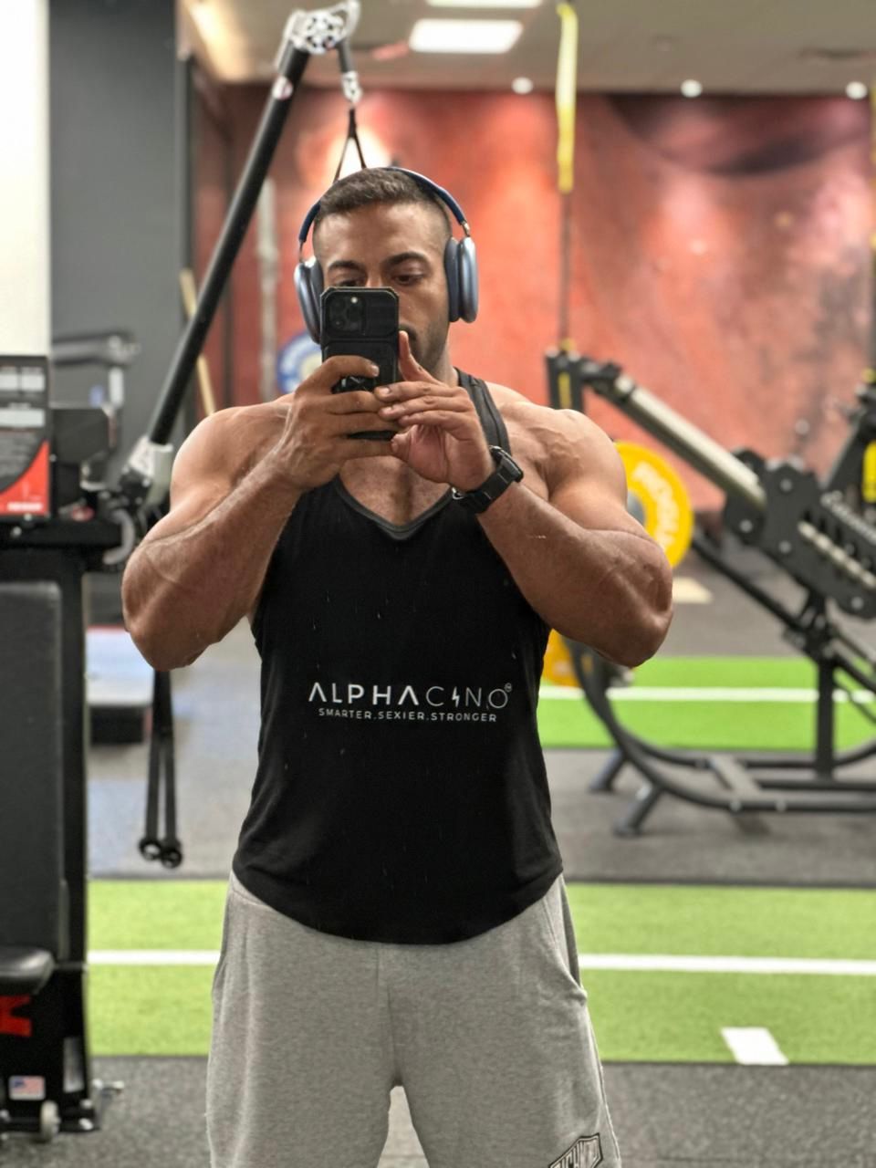 Man in an Alphacino tank top taking a mirror selfie in a gym between sets